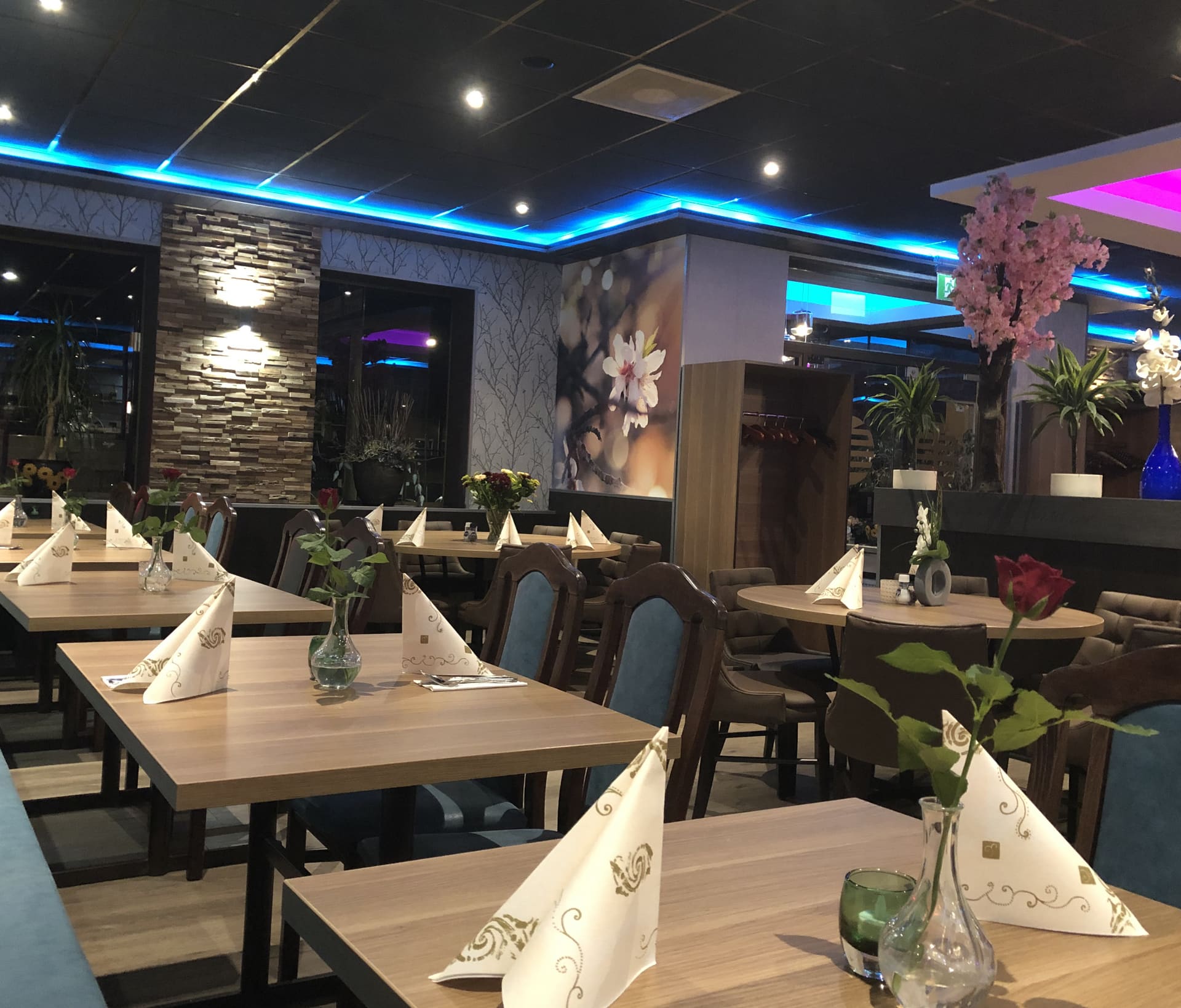 De eetzaal van Sozing met gedekte tafels, servetten en bloemen onder blauwe plafondverlichting.